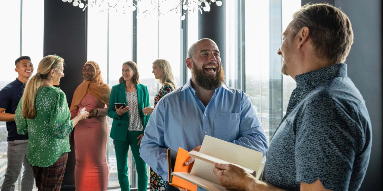 Two white men both wearing blue shirts chat standing in an office setting with five women and a man chatting in the background